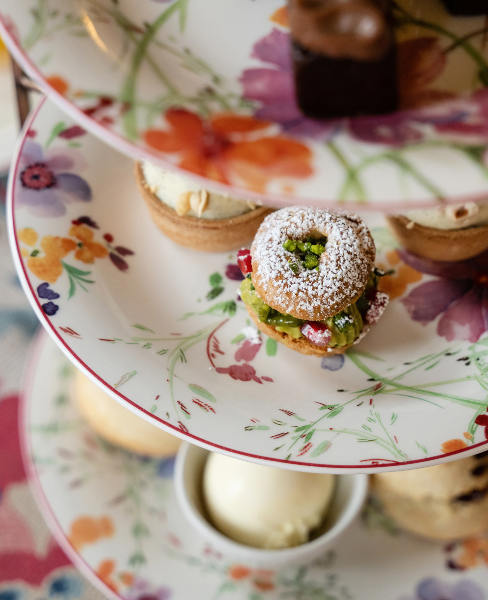 A close-up shot of a floral-patterned tiered serving stand filled with various afternoon tea delicacies at the Cromlix Hotel, including a powdered sugar-dusted pastry with green filling and red berries on the top tier, a tart and a dark chocolate treat on the middle tier, and a glimpse of other pastries and a small white bowl (possibly containing cream or curd) on the lower tier.