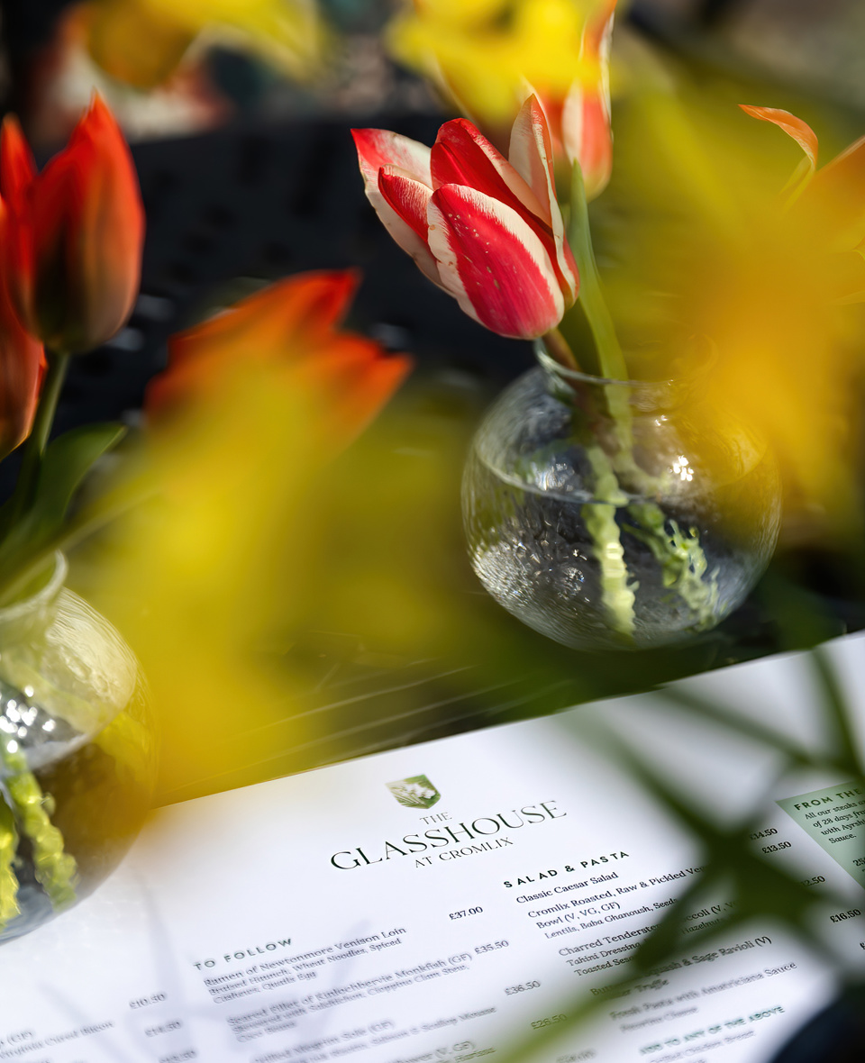 A softly focused image showing a menu from The Glasshouse at Cromlix, partially visible on a dark surface, with two small, clear glass vases each holding a few red and white tulips. The background is blurred with yellow and green hues, suggesting an outdoor or bright setting.