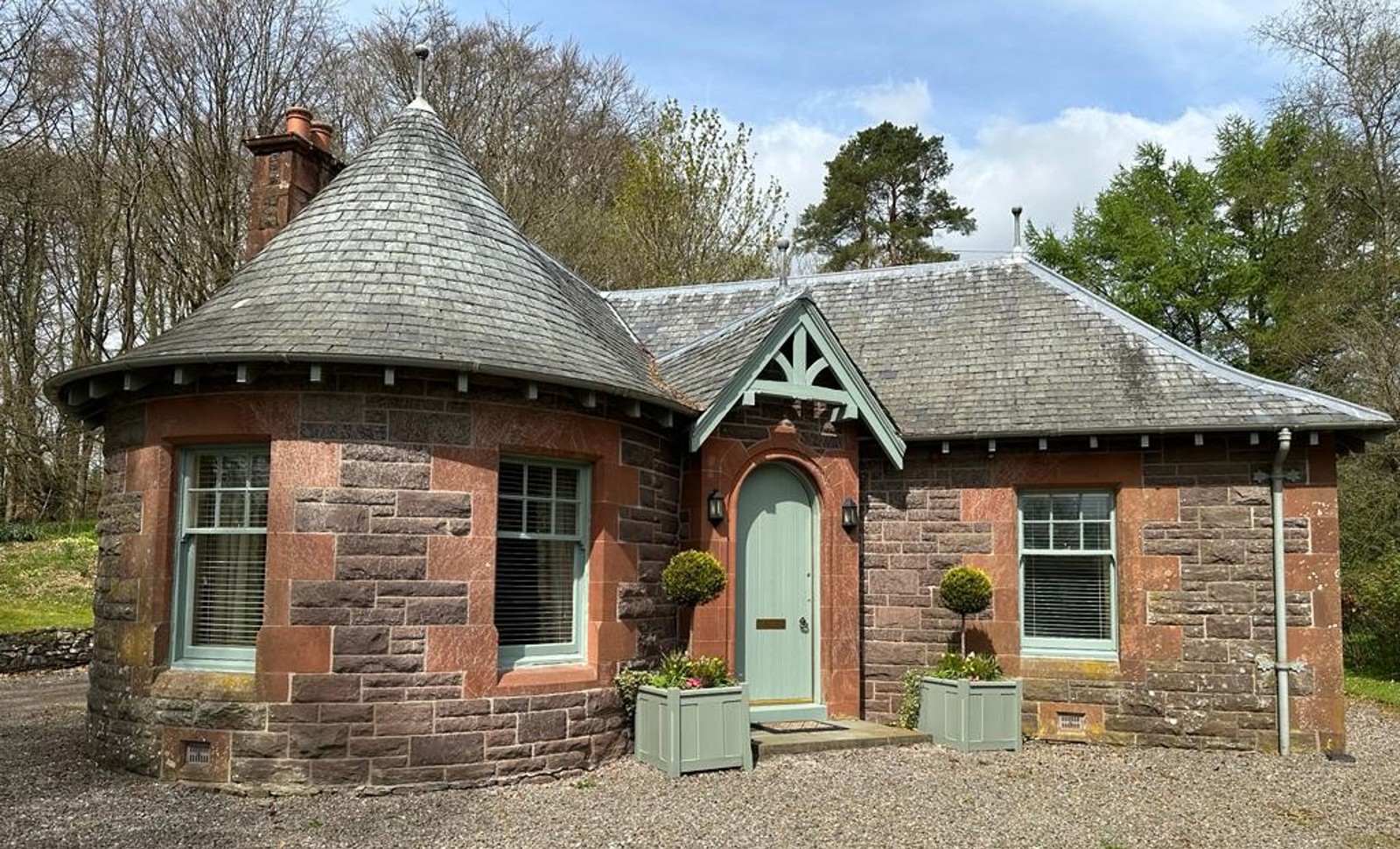 Charming exterior of The Laurel Gate Lodge at Cromlix House in Dunblane, with its red stone walls, grey slate roof, a distinctive turret, a green front door, and potted plants.