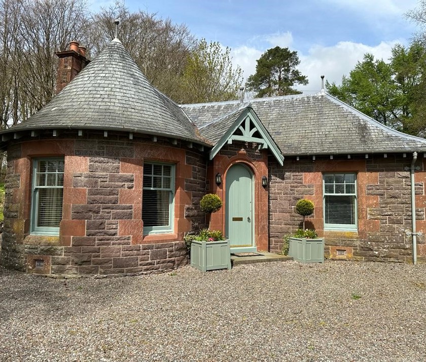 Charming exterior of The Laurel Gate Lodge at Cromlix House in Dunblane, with its red stone walls, grey slate roof, a distinctive turret, a green front door, and potted plants.